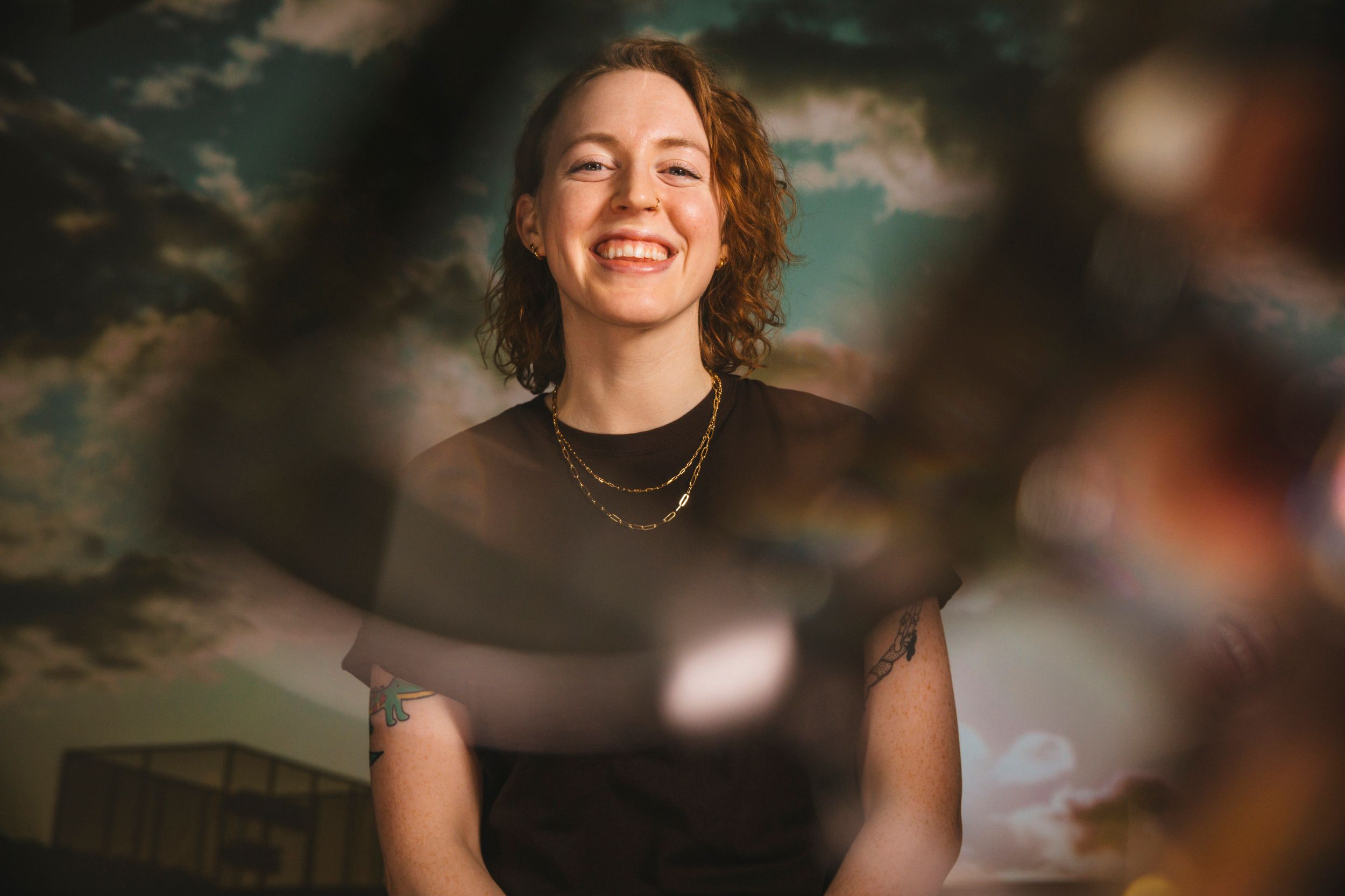 Nora, a white person with curly red hair and wearing a brown t-shirt, is smiling against a cloudy backdrop. An artful camera blur appears on top of the image.