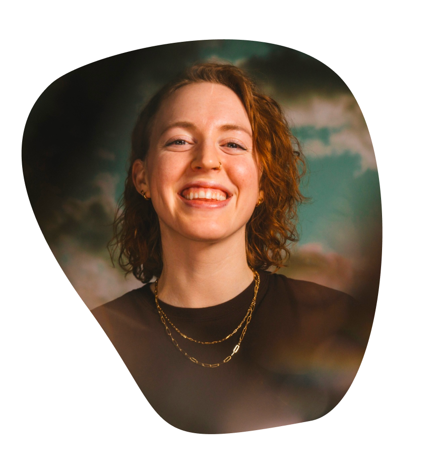 Nora, a white person with red curly hair, is smiling at the camera. The photo is cut in a playful shape, like a doodle on paper.