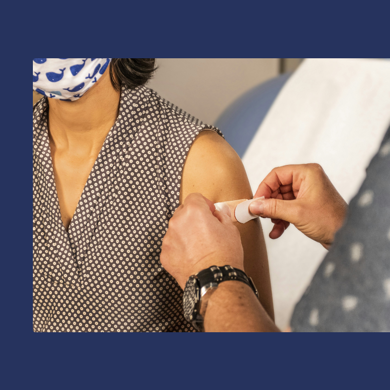 A person is seated and wearing a medical mask while another person places a small bandage on their upper arm.