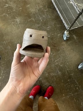 A clay-covered hand holds clay thrown and carved into a votive holder with a silly frowny face. The ceramicist's clay-covered brown pants and red clogs are visible.