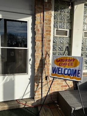 An easel next to a patio door holds a handpainted sign in the style of Chicago grocery stores that says SATURDAY COFFEE WELCOME.