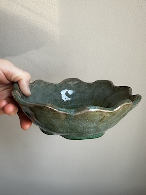 A hand holds out a greenish-blue handbuilt ceramic bowl with a scalloped top edge. The shiny glaze seems to drip off the bottom of the bowl.