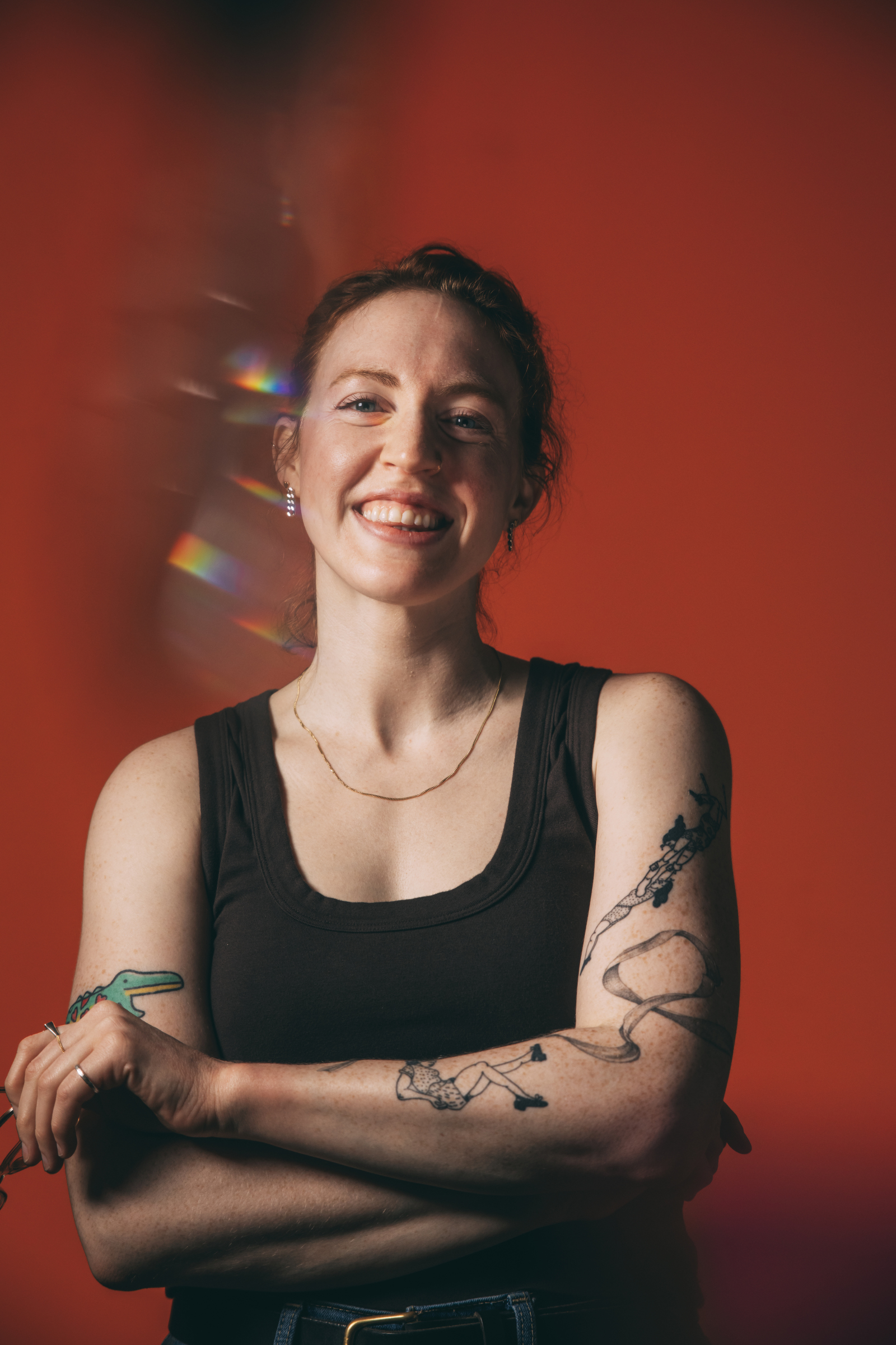 Nora Hanlon, a white person with red curly hair tied back in a bun, faces the camera smiling with her arms crossed in a relaxed position. She's wearing a tank top and has playful tattoos visible on her arms.